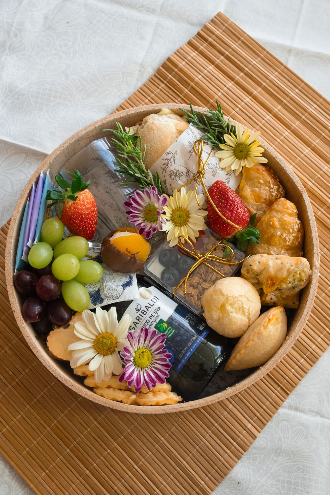 Cesta Amor que Alegra em base redonda de madeira com uvas, morangos, salgados folhados, flores e suco integral.